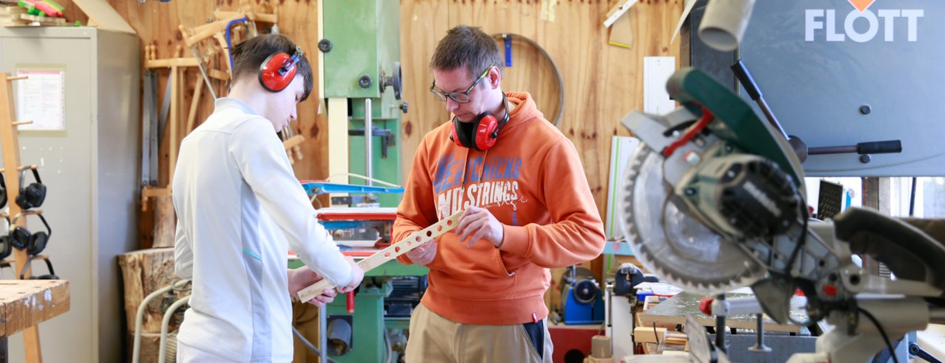 ein junger Mann und sein Anleiter tragen beide roten Gehörschutz und arbeiten an einem Werkstück aus Holz arbeiten