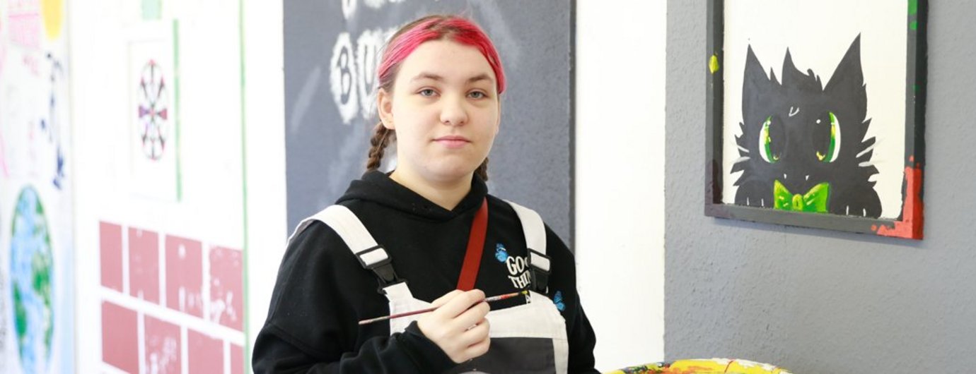 Eine junge Frau mit roten Haaren und weißer Latzhose steht mit Pinsel und farbe vor einer Zeichnung mit Katze
