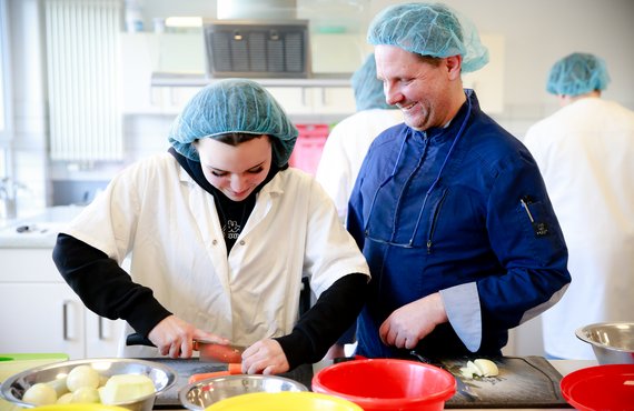 eine junge Frau bekommt von einem Mann das Schneiden von Gemüse erklärt. Beide tragen Schutzhauben.